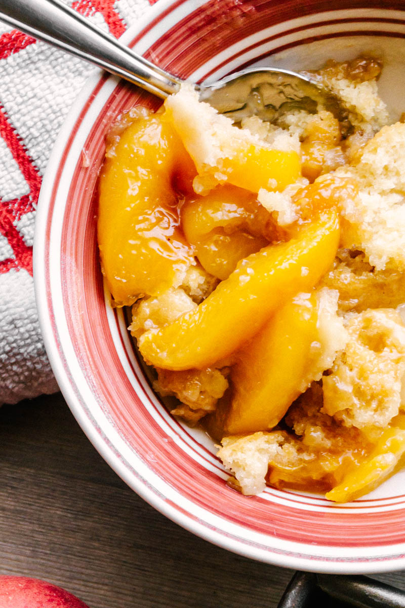 peach cobbler in a red bowl with a spoon and peaches