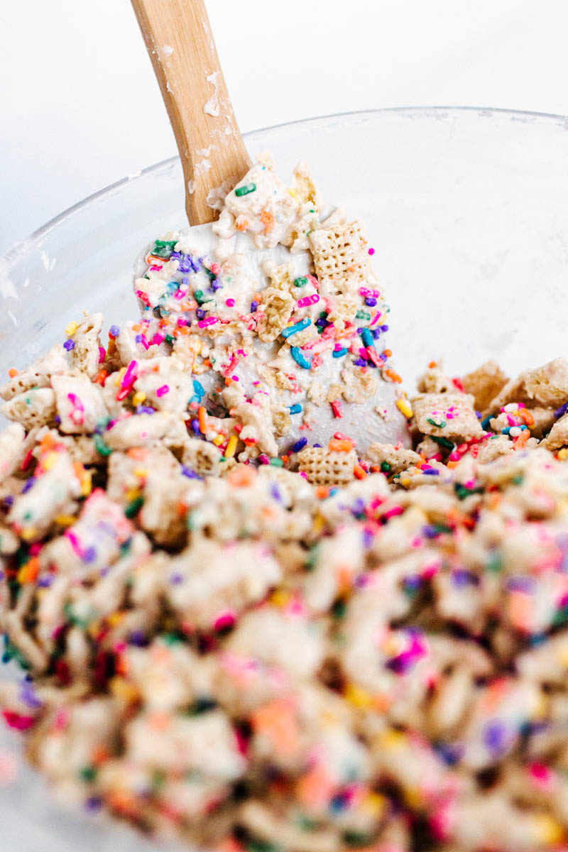 cake batter muddy buddies being mixed in a bowl with a spatula coated in sprinkles