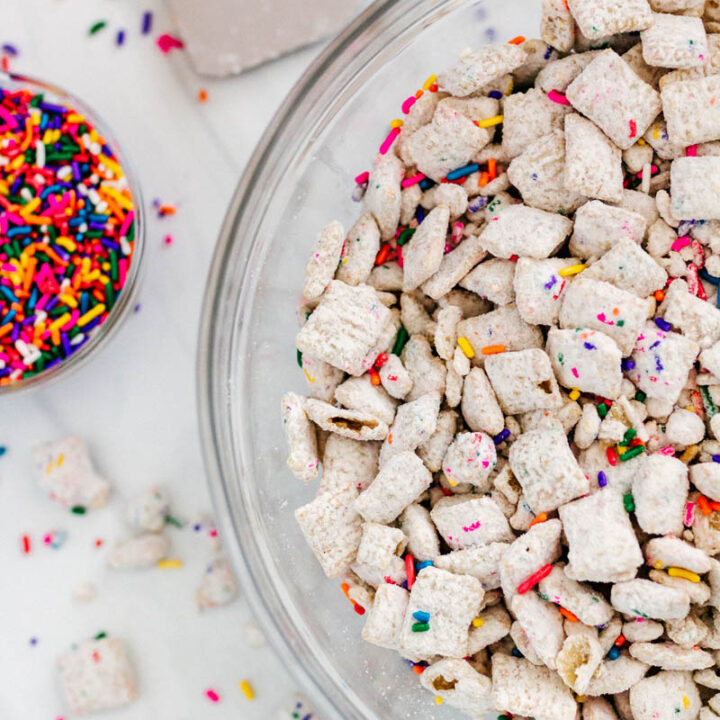 cake batter muddy buddies with sprinkles in a bowl, with sprinkles and a spatula next to it