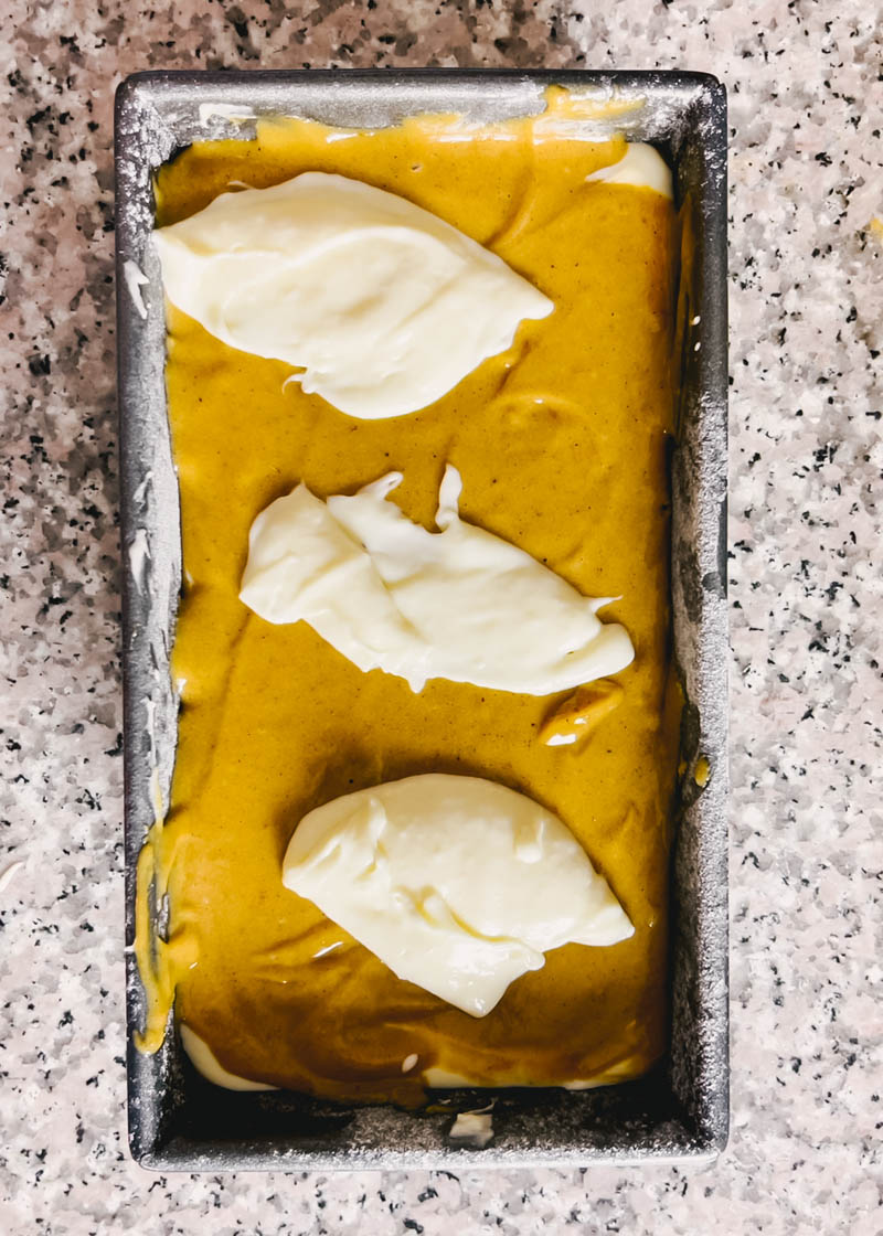 pumpkin bread batter layering in a loaf pan with 3 large dollops of cream cheese on top