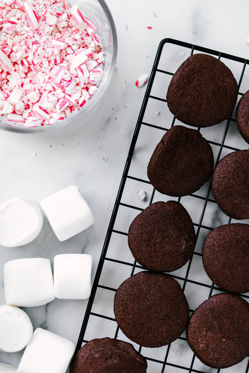 chocolate peppermint cookies on a wire rack with marshmallows and crushed candy canes nearby