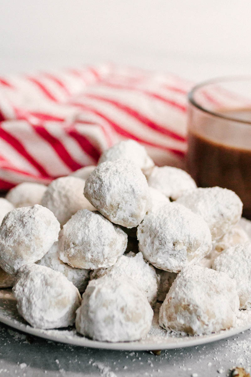 snowball cookies stacked on a plate with hot chocolate and a towel in the background