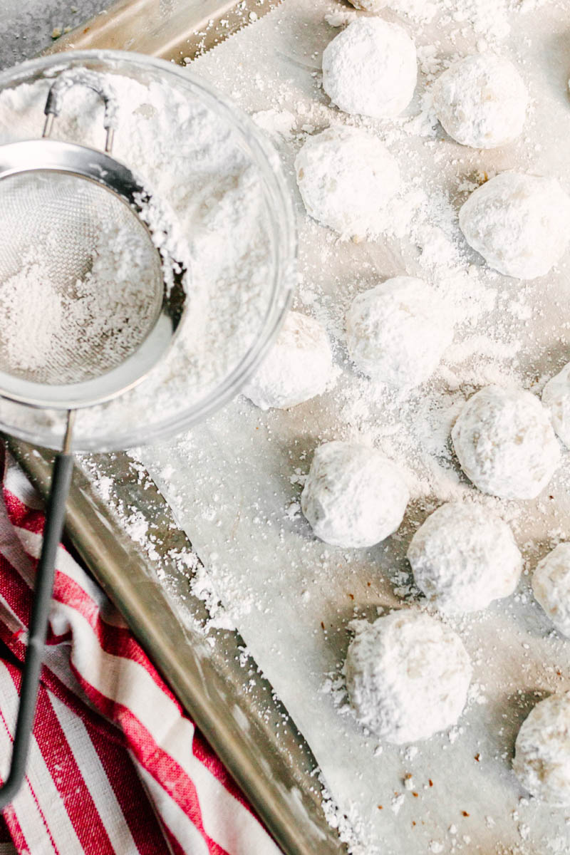 snowball cookies on a baking sheet getting dusted with powdered sugar
